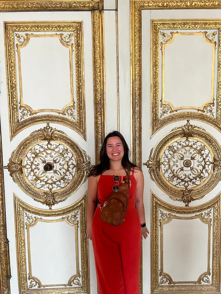 Natalie standing in front of white doors with golden accents on them. She is smiling, wearing red overalls with her hands by her side. She has a brown cross-body bag across her chest.