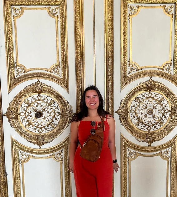 Natalie standing in front of white doors with golden accents on them. She is smiling, wearing red overalls with her hands by her side. She has a brown cross-body bag across her chest.