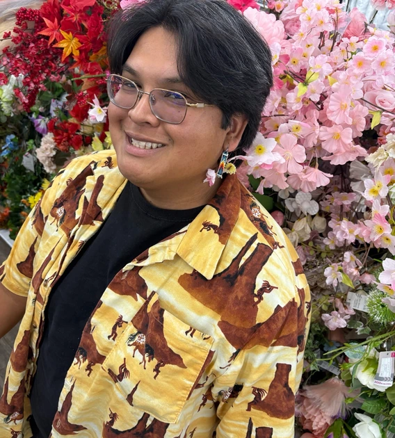 Ivan standing in front of many different colored flowers, smiling up at the camera. He is wearing an open-buttoned shirt with a black t-shirt underneath.