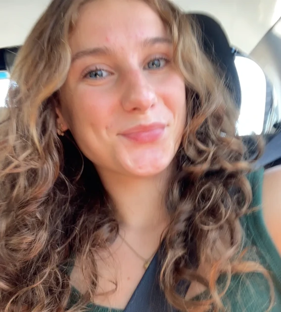 Alex smiling at the camera in a selfie. She is sitting in a car, head slightly tilted with her curly hair pulled over her shoulders.